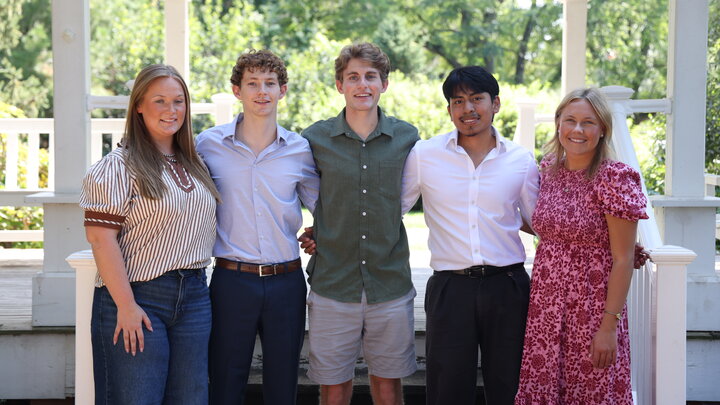 Group of five people smiling outdoors on a wooden porch with greenery in the background.