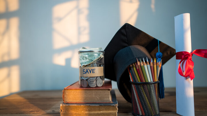 Graduation cap, diploma, jar labeled "save," pencils, and books on a wooden table with sunlight.