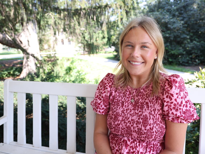 Smiling woman in a pink floral dress sits on a white bench outdoors in lush greenery.