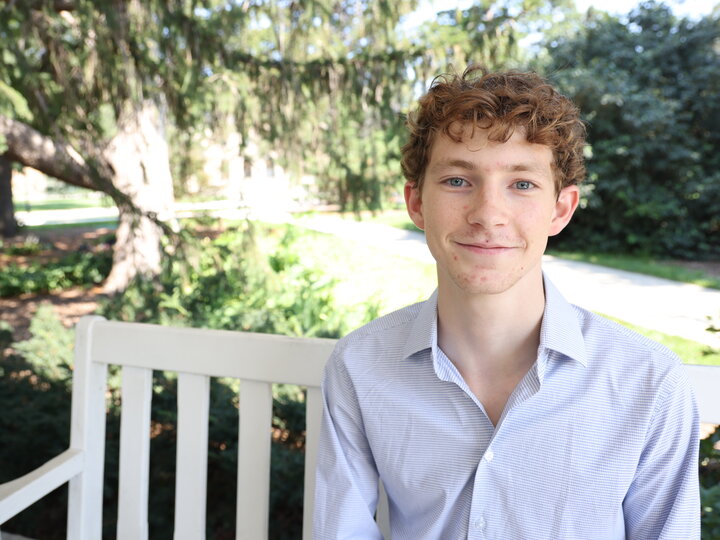 Smiling person sitting on a white bench in a sunlit park with green trees in the background.