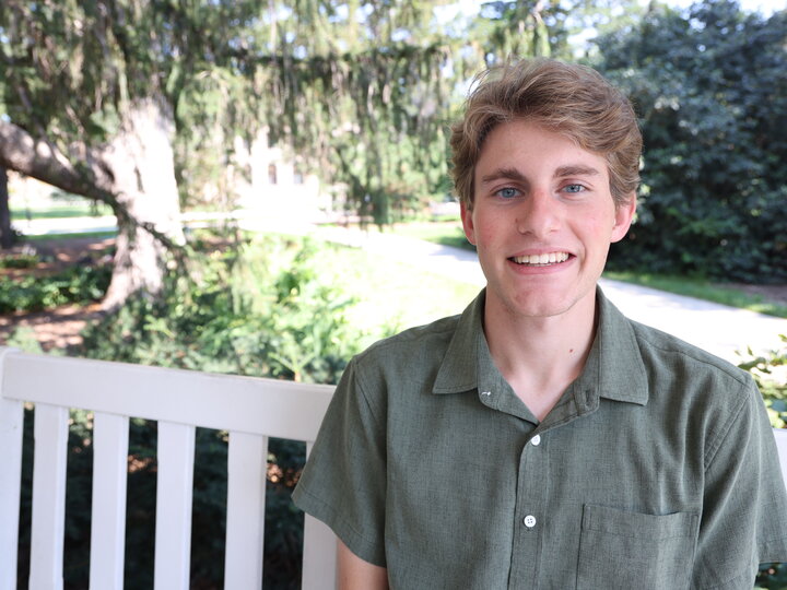 Smiling man sitting on a white bench outdoors, surrounded by trees and greenery.