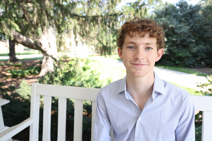 Smiling person sitting on a white bench in a sunlit park with green trees in the background.