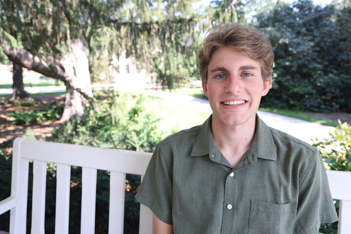 Smiling man sitting on a white bench outdoors, surrounded by trees and greenery.